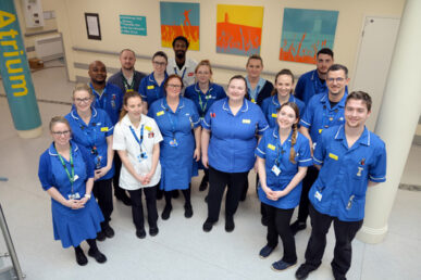 A group of fifteen Smiley Somerset FT colleagues, some in blue uniform with black trousers, some with white tops and some with blue dress uniforms. Standing shoulder to shoulder in the Beacon Centre Atrium.