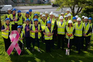 Breast care fundraisers in blue hard hats, breast care team and builders in white hard hats stand wearing hi vis jackets outside Yeovil Hospital at the site the new breast care unit will be built. The group are gathered, looking towards the camera with a road in the background. At the front, the breast care consultant is posed with a shiny spade and a fund raiser in a wheelchair in the front of the photo holds a large pink ribbon cut out image.