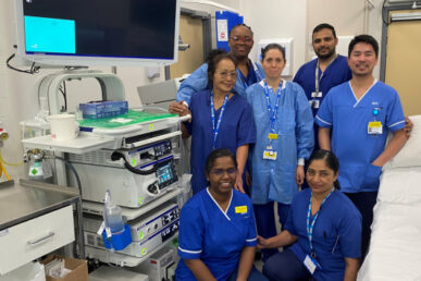 A group of six Somerset NHS FT colleagues in blue scrubs looking happy to be in the light and bright clinical room. There is a bed on one side of the group and an Endoscopy machine on the left hand side with a computer screen on top.