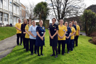 Group of lovely smiling uniformed colleagues stood on the grass in a line with Yeovil hospital in the background. there are yellow, blue and white uniforms.