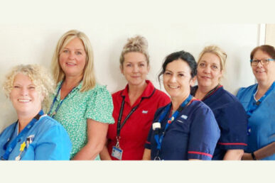 Our homeless and rough sleepers service is a community-based team and photographed are six members of the Somerset NHS Foundation Trust team stood in a line with their bodies facing the left of the image and smiling faces to the front. The colleagues at each end are wearing light blue uniform blouses. Two colleagues to the right of the centre are wearing navy with red trim nursing uniforms and in the centre a nurse is wearing a red with navy trim uniform. The colleague to the left of the sentre is not in uniform and wears a green and white dress.