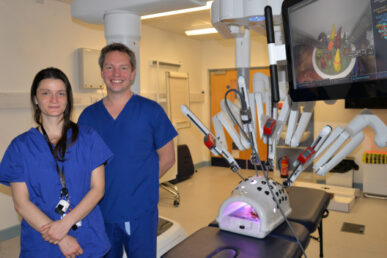 Mr Richard Bamford, one of our colorectal surgeons and another woman colleague, both in blue theatre scrubs, stood in front of the robot. The robot has lots of different arms coming from the main body. They are demonstrating how the different arms work on a dummy,