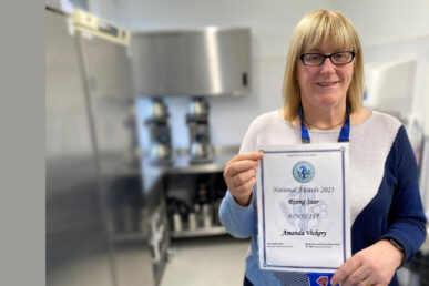 Our catering team leader at Musgrove Park Hospital, Amanda Vickery, is photographed from the waist up holding her catering certificate showing she reached the final of this year’s Hospital Caterers’ Association awards. She has blonde shoulder length hair, a fringe and is wearing glasses. The background is a blurred professional kitchen setting.