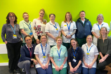 Somerset’s Discharge to Assess service team members are photographed in two rows with six colleagues crouched down at the front and a further eight colleagues standing behind them against a bright green backdrop. Six of the colleagues are wearing smart/casual day clothes and the rest are in uniform with a mixture of light green tunic tops and white tunics with navy blue trim, representing the different clinical and non clinical roles within the team.