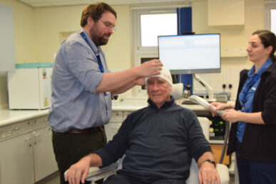 Two Somerset FT colleagues in a clinical room, stood either side of a patient sat on a medical chair. The patient has a headband covering his head which has wires coming out attached to a computer. There is a computer screen in the background monitoring results.