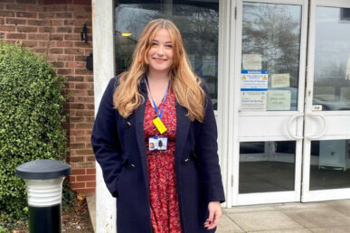 Sophie, wearing a floral red dress, with a blue parker jacket is stood in front of double white doors which is the entrance to Lower Bracken house. On the left hand side is some green tall buses with a four foot pole with a night light on the top.