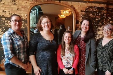 Five advance care planning colleagues all stood in smart casual clothing stood in a floor to ceiling mirror which is against a light brick wall.