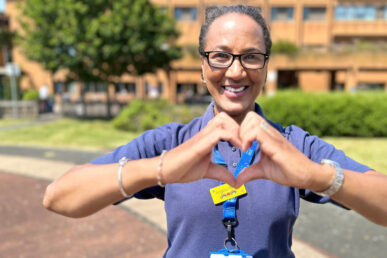 Angela Reece, a specialist nurse for children who is stood on the helicoptor landing pad in front of the main building at Musgrove. She is got her arms at her chest height, creating a heart shaped hand gesture.