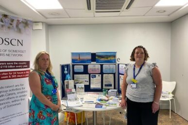 Two colleagues one in a blue flowery dress and the other in a white top with a black skirt, stood in front of a stand with lots of information such as leaflets, booklets, and posters, about how we are supporting carers across the county. They are in a white well lit room, with a wooden floor.