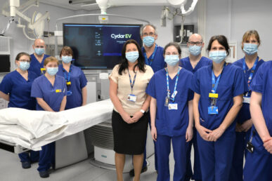Group of eleven Somerset NHS FT colleagues, some in blue uniform, and a consultant in non uniform. Stood in a clean white clinical room next to a movable bed with a large computer screen in the background which show results.