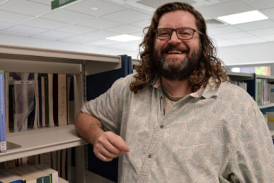 Man wearing a light patterned shirt smiling and leaning on a book shelf.