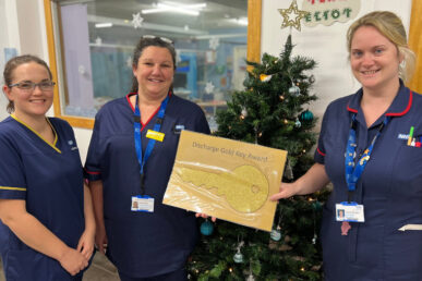 Three nurses at Musgrove Park Hospital holding a discharge key award. They are stood next to a Christmas tree on a hospital ward.
